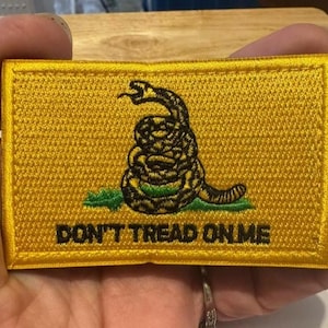 May include: A yellow embroidered patch featuring a coiled rattlesnake above green grass. The patch has a black border and the words "DON'T TREAD ON ME" in black block letters.