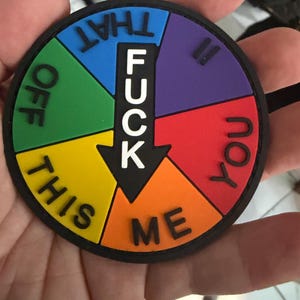 May include: A round, colorful patch with a black border. The patch is divided into pie-shaped sections of yellow, green, blue, purple, red, and orange. The word "FUCK" is in white, with an arrow pointing to the orange section, and other words are around the circle.
