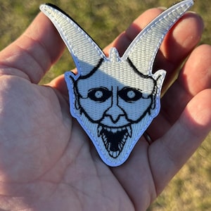 May include: Embroidered patch featuring a white demonic face with black outlines. The patch has a detailed design of a horned head with a wide, toothy grin. The patch is held in a hand against a blurred green background.