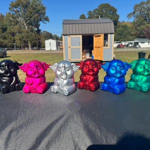 May include: A collection of six colorful elephant figurines in black, pink, silver, red, blue, and green. The figurines are sitting on a dark surface, with a background of a house and trees.