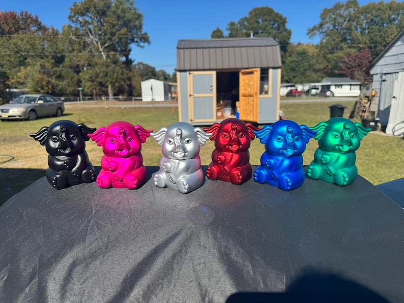 May include: A collection of six colorful elephant figurines in black, pink, silver, red, blue, and green. The figurines are sitting on a dark surface, with a background of a house and trees.