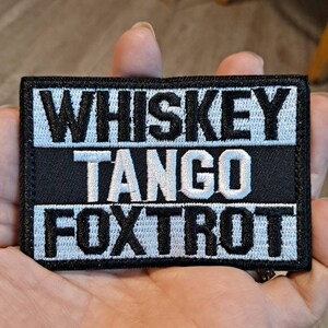 May include: Black and white embroidered patch with the words "WHISKEY TANGO FOXTROT" in bold, capital letters. The patch has a rectangular shape with a black background and white stitching.