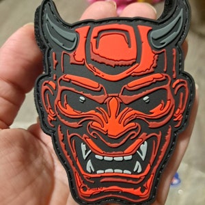 May include: A black and red patch featuring a stylized Oni mask design. The mask has prominent horns, sharp teeth, and intricate details. The patch is likely made of rubber or a similar material, with a hook and loop backing.
