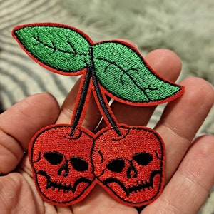 May include: Two red cherries with black skull faces and green leaves. The cherries are embroidered on a fabric patch.