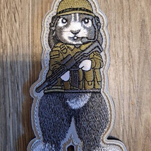 May include: A gray embroidered patch of a cartoon rodent wearing a military helmet and uniform, holding a rifle.