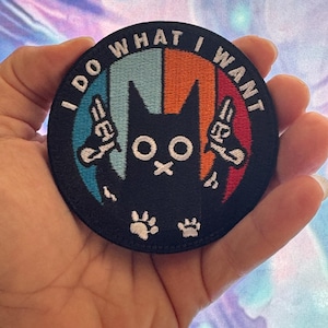 May include: Round black patch with the text "I DO WHAT I WANT" in white. The patch features a black cat with white eyes and paws, holding two guns. The background is divided into blue, orange, and red sections.
