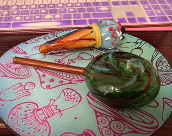 May include: Two glass pipes on a blue and pink patterned surface. One pipe is green and shaped like a swirl. The other pipe is orange, yellow, and blue and shaped like an ice cream cone.