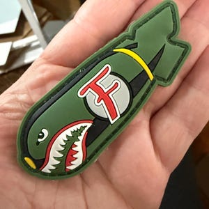 May include: A green patch in the shape of a bomb, featuring a shark mouth design with white teeth and red accents. The patch includes a yellow stripe and a circular emblem with the letter "F".