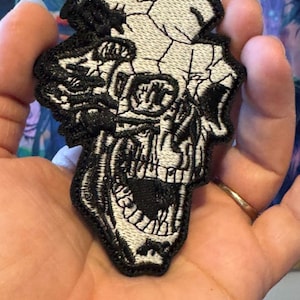 May include: Embroidered patch featuring a detailed, black and white skull design. The skull has a cracked appearance on the top and an open mouth. The patch is likely for clothing or accessories.