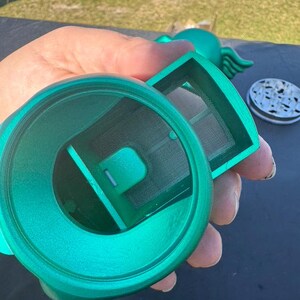 May include: A teal-colored tea infuser with a mesh filter and a lid. The infuser is held in a hand, showcasing its design and components. The lid has a speckled pattern.