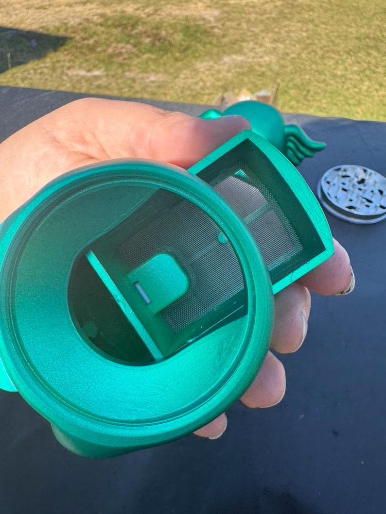 May include: A teal-colored tea infuser with a mesh filter and a lid. The infuser is held in a hand, showcasing its design and components. The lid has a speckled pattern.