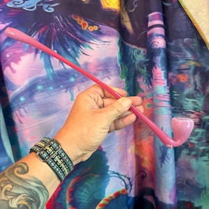 May include: A pink glass pipe with a curved stem and bowl. The pipe is held against a colorful, patterned background. The pipe's design is simple and elegant, with a smooth, glossy finish.