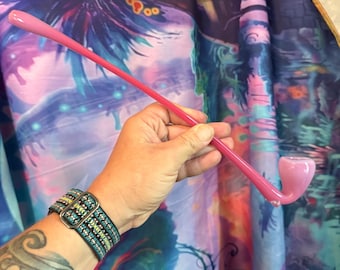 May include: A pink glass pipe with a curved stem and bowl. The pipe is held against a colourful, patterned background. The pipe's design is simple and elegant, with a smooth, glossy finish.