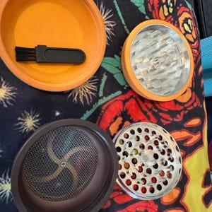 May include: An orange herb grinder with a brush, and multiple components. The grinder has a silver metal grinding surface and a brown mesh screen. The components are arranged on a colorful floral patterned surface.