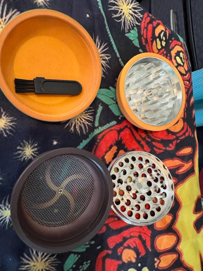 May include: An orange herb grinder with a brush, and multiple components. The grinder has a silver metal grinding surface and a brown mesh screen. The components are arranged on a colorful floral patterned surface.