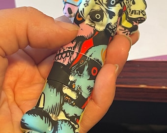 May include: A colorful silicone pipe with a skull design. The pipe is decorated with a variety of skulls and other graphic elements in red, blue, yellow, black, and white.