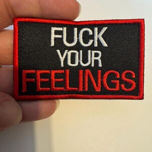 May include: Black rectangular patch with red border and text "FUCK YOUR FEELINGS" in white and red thread. The patch is held by a hand.