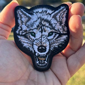 May include: Embroidered patch depicting a wolf's head with a snarling expression. The wolf has white and grey fur, green eyes, and a black border. The patch is designed for application on clothing or accessories.