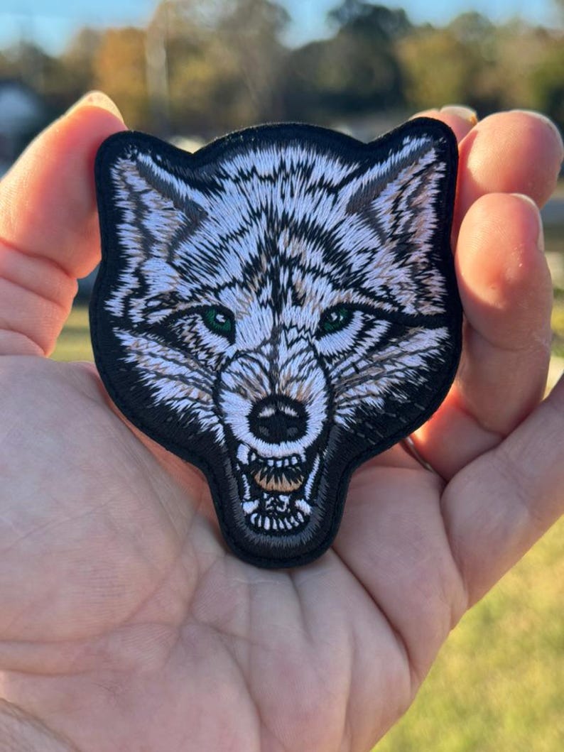 May include: Embroidered patch depicting a wolf's head with a snarling expression. The wolf has white and grey fur, green eyes, and a black border. The patch is designed for application on clothing or accessories.
