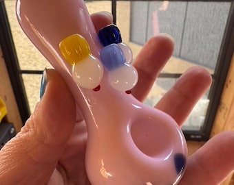 May include: A pink glass pipe with a round bowl and a small handle. The pipe is decorated with colorful glass accents, including blue, yellow, and white.