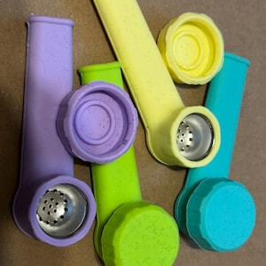 May include: A collection of brightly coloured silicone pipes in purple, green, yellow, and teal. Each pipe has a metal screen and a matching silicone cap. The pipes are displayed on a neutral-toned surface.