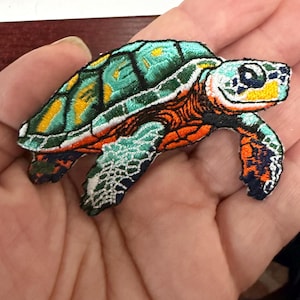 May include: Embroidered sea turtle patch featuring a detailed design with teal, orange, yellow, and black thread. The turtle's shell has a patterned design, and the edges are neatly stitched. This patch is suitable for adding to clothing or accessories.