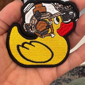 May include: Embroidered patch featuring a yellow rubber duck with a black outline and a red beak. The duck is wearing a detailed military-style helmet and gear, with brown, white, and black accents. The patch has a black border.