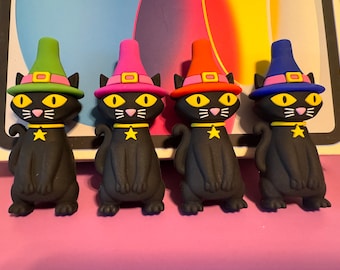 May include: Four black witch cat figurines with different colored hats. The hats are green, pink, orange, and blue. Each cat has a yellow star on its collar.