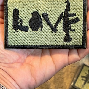 May include: A green embroidered patch with the word "LOVE" spelled out using the silhouettes of a gun, a grenade, a knife, and a rifle.