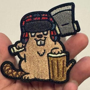 May include: Embroidered patch featuring a cartoon beaver wearing a plaid hat and holding an axe and a mug of beer. The beaver is brown with white teeth and a black nose. The axe is gray, and the mug is tan.