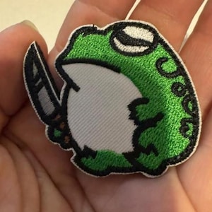 May include: Embroidered patch featuring a green frog holding a knife. The frog has a white belly, black eyes, and a green head. The patch has a black border and is likely for clothing or accessories.