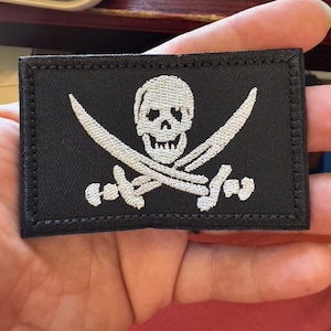 May include: Black rectangular patch featuring a white skull and crossbones design. The skull is centered above two crossed swords. The patch has a black border and is likely an embroidered emblem or patch.