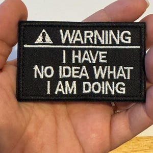 May include: Black rectangular patch with white embroidered text that reads "WARNING I HAVE NO IDEA WHAT I AM DOING". The patch has a white triangle with an exclamation point symbol above the word "WARNING".
