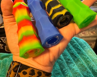 May include: Four silicone smoking pipes in different colors and patterns. The pipes are in a hand, and the hand is wearing a leopard print watch band.