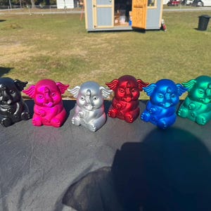 May include: A row of six colorful elephant figurines. The elephants are in various metallic colors: black, pink, silver, red, blue, and green. Each figurine has a detailed trunk, ears, and a third eye.