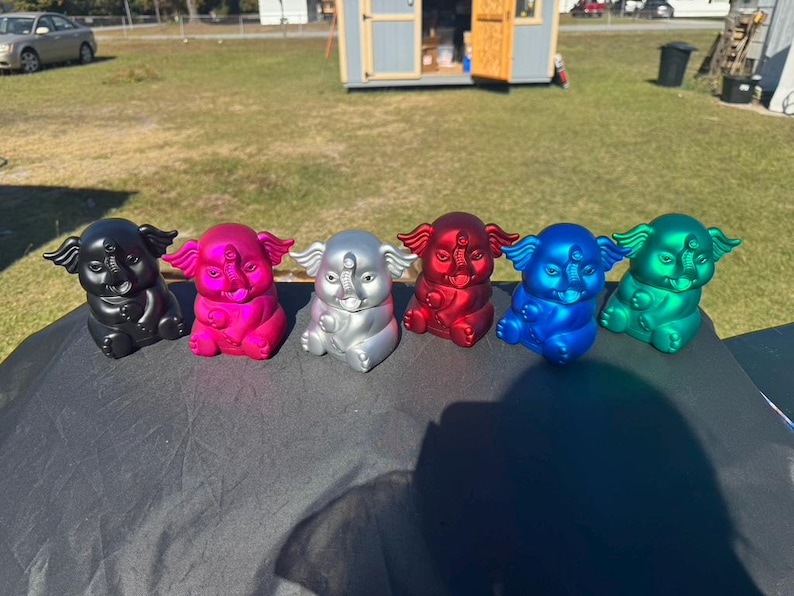 May include: A row of six colorful elephant figurines. The elephants are in various metallic colors: black, pink, silver, red, blue, and green. Each figurine has a detailed trunk, ears, and a third eye.