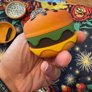 May include: A novelty burger-shaped grinder with orange, green, brown, and yellow sections. The top bun has black seed details. Additional orange and silver grinders and a small tray are visible in the background.