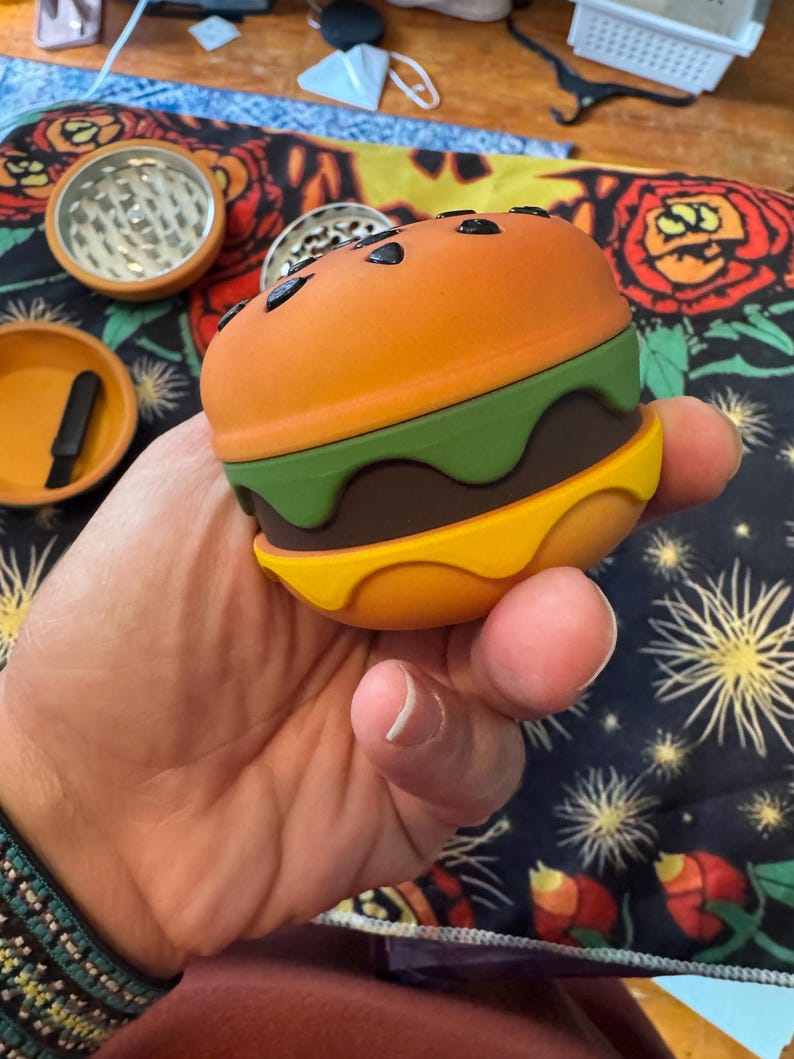 May include: A novelty burger-shaped grinder with orange, green, brown, and yellow sections. The top bun has black seed details. Additional orange and silver grinders and a small tray are visible in the background.