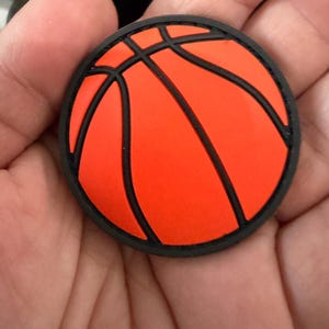 May include: A round, orange basketball charm with black lines and a black border. The charm is held in a hand, showcasing the vibrant colors and design. This accessory is perfect for sports enthusiasts.