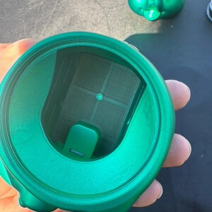May include: A close-up of a green, elephant-shaped container with a mesh filter inside. The container is made of a shiny, metallic material. The container is held in a person's hand.