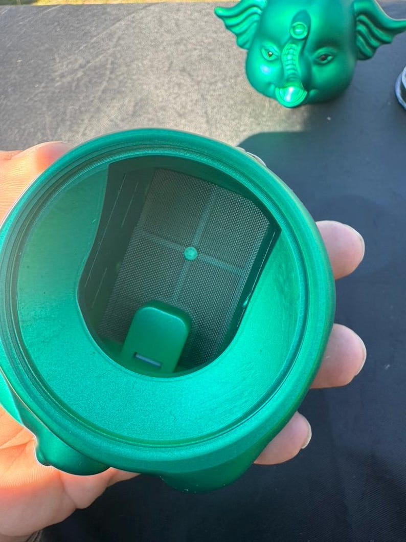 May include: A close-up of a green, elephant-shaped container with a mesh filter inside. The container is made of a shiny, metallic material. The container is held in a person's hand.