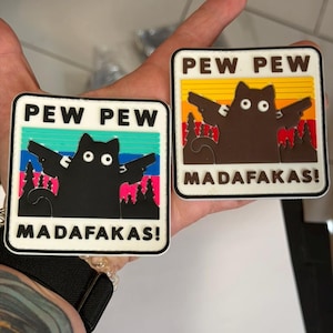 May include: Two square patches with a black cat silhouette holding guns, the text "PEW PEW MADAFAKAS!" is displayed. One patch has a colorful background, the other has a brown cat with a sunset background.