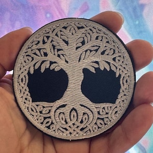 May include: Round embroidered patch featuring a white tree of life design on a black background. The tree's branches and roots are intricately detailed. The patch has a white border and is held in a hand.