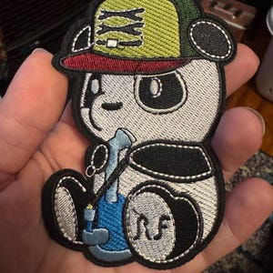 May include: An embroidered iron-on patch featuring a panda wearing a green and yellow baseball cap with a red brim, holding a blue bong. The panda is wearing a black and white shirt and has a black and white face with black eyes. The patch has the letters "NS" embroidered on the bottom right corner.