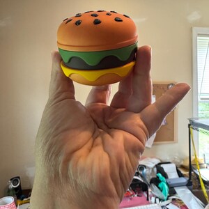 May include: A novelty burger-shaped container with an orange top bun dotted with black seeds, a green lettuce layer, a brown patty, and a yellow cheese bottom bun. The container is held in a hand.