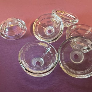 May include: Six clear glass bottle stoppers with a small hole in the center of each. The stoppers are on a purple surface.