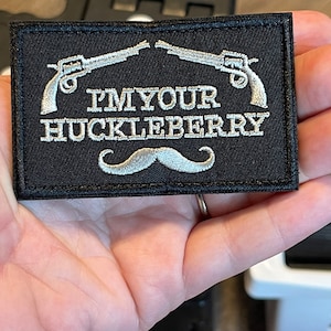 May include: A black embroidered patch with white stitching. The patch features two crossed pistols, a mustache, and the text "I'M YOUR HUCKLEBERRY".