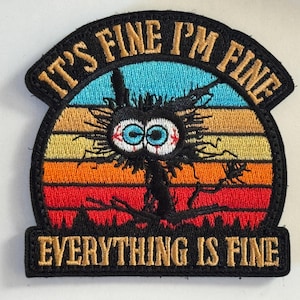 May include: Embroidered patch with the phrase "IT'S FINE I'M FINE EVERYTHING IS FINE". The patch features a distressed cat design with large, bloodshot eyes, set against a sunset gradient of blue, yellow, orange, and red.