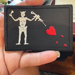May include: Black rectangular patch with a white embroidered skeleton holding a rifle and a glass. A red heart and three red dots are embroidered on the right side. The patch has a black border.