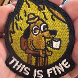 May include: Embroidered patch featuring a cartoon dog sitting in a chair, holding a mug, with the text "THIS IS FINE" in white letters. The dog is brown with a yellow background and black border.